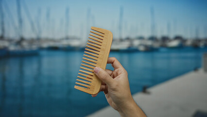 Man holding wooden comb by the seaside with blurred boats in the background at a port, highlighting...