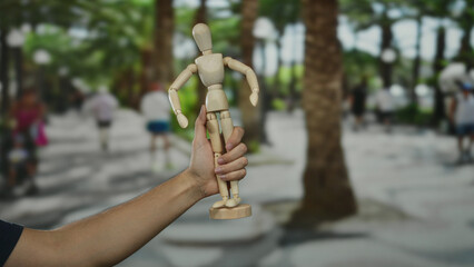 Hand holding wooden mannequin on a city street with blurred figures in the background depicting...