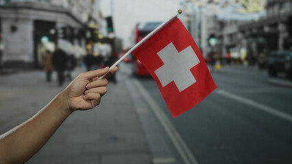 Male hand holding swiss flag in bustling city street with blurred red bus in background symbolizes...