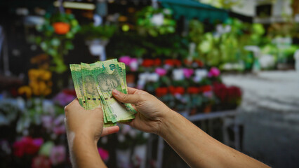 Man holding south african rand in outdoor flower shop with vibrant blooms, emphasizing currency transaction and financial exchange in an exterior setting.