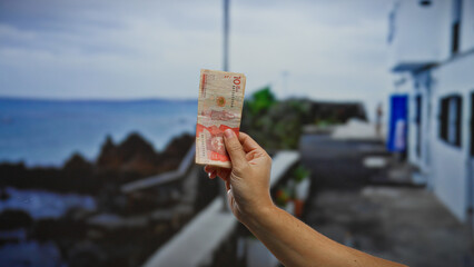 Hand holding colombian pesos by a seaside town street, highlighting vacation spending with local...