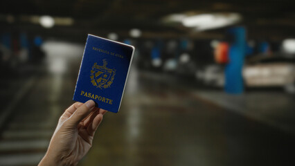 Hand holding a cuban passport indoors in a dimly lit parking garage with cars visible in the background, suggesting travel or identity verification by a man.