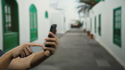 Man engaged with smartphone on a sunny city street lined with white buildings and green windows, capturing a moment of digital interaction in an outdoor urban environment.