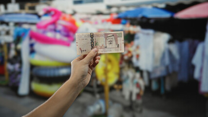 Man holding saudi riyal banknote outdoors in a vibrant souvenir shop setting with colorful items displayed, depicting a lively marketplace atmosphere.