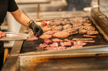 Chef placing raw meat onto a hot grill in a professional kitchen. A variety of meats are sizzling over open flame with smoke rising in a busy cooking environment.