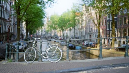 Obraz premium Bicycle standing defocused on an amsterdam canal bridge with blurred buildings, trees, and water in the background under bright sunlight.
