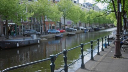 Fototapeta premium Blurred view of amsterdam canals with trees, buildings, and boats reflecting in the water creating a serene, peaceful atmosphere with a defocused, bokeh effect.