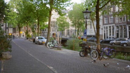 Defocused view of a scenic street along amsterdam's canals featuring parked bicycles, lamp posts, and classic dutch architecture, with spring foliage creating a verdant cityscape. © Krakenimages.com