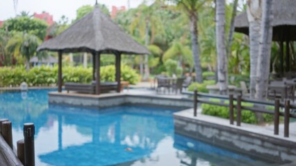 Blurred tropical resort with defocused luxury pool area, lush greenery, and thatched-roof gazebo...