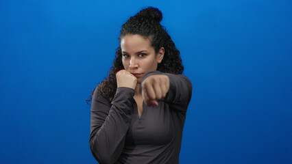 Young hispanic woman poses with fists raised against an isolated blue wall background exuding...