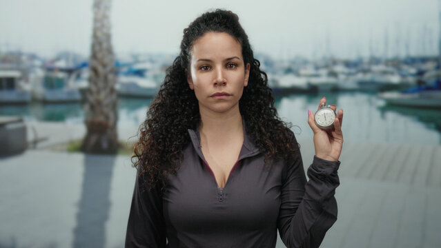 Woman holding stopwatch at seaside port, wearing athletic attire, showcasing determination and focus with boats and sea in the background under a cloudy sky.