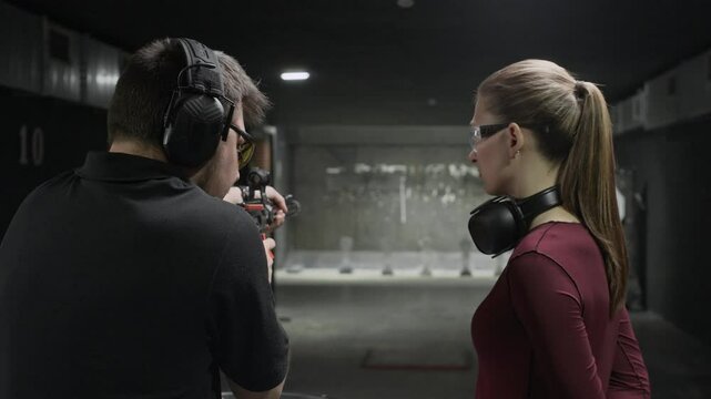 Back view of young female Caucasian trainee wearing goggles listening to male instructor teaching to hold rifle at indoor range