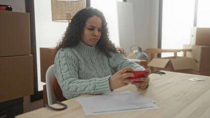 Woman unpacking in new apartment surrounded by boxes checks phone at wooden table with paperwork...