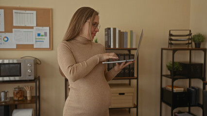 Pregnant woman working on laptop in office setting, surrounded by books, binders, and charts, illustrating modern professional lifestyle.