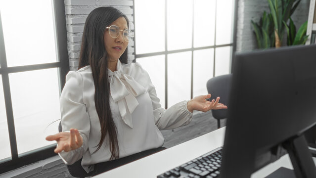 Woman gesturing in modern office with computer, wearing glasses and elegant white blouse, suggesting confusion or questioning during a work-related task, indoors.