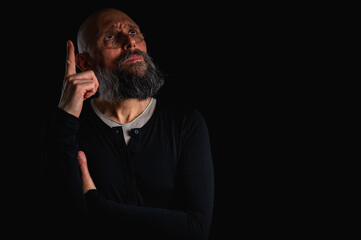 Thoughtful bald man with beard and glasses looking up, touching temple, deep in thought, isolated on black background. Concept of ideas, planning, decision-making