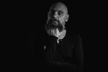Black and white portrait, pensive bald man with beard and glasses looking up, touching temple, deep in thought, isolated on black background. Concept of ideas, planning, decision making