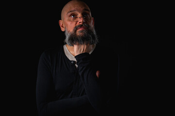 Thoughtful bald man with beard and glasses looking up, touching temple, deep in thought, isolated on black background. Concept of ideas, planning, decision-making