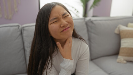 Woman with sore throat sitting on a couch in a modern living room, expressing discomfort indoors, showcasing an asian female in a home setting.