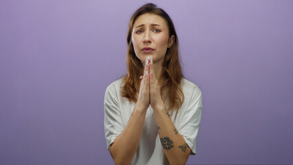 Woman with tattoos in a white shirt pleading with hands together in front of a purple background,...