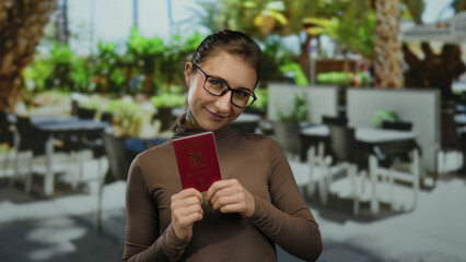 Young woman holding finnish passport at outdoor restaurant terrace, smiling warmly in a casual setting, highlighting finland's travel documentation.