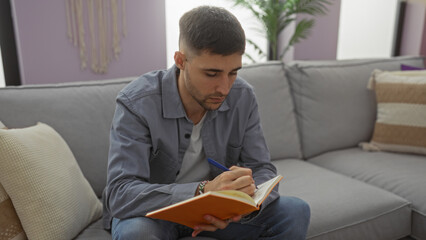 Young man writing in notebook while sitting on cozy sofa in modern living room surrounded by soft pillows emphasizing a comfortable and creative home environment.