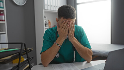 Tired hispanic male doctor in green scrubs rests face in hands at clinic desk with medical documents and laptop, under bright indoor lighting during a busy workday.