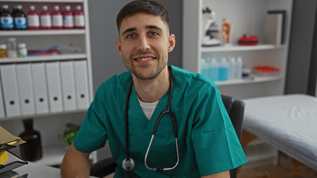 Young man in clinic wearing scrubs smiling confidently in hospital room setting with medical equipment showcasing professional healthcare environment