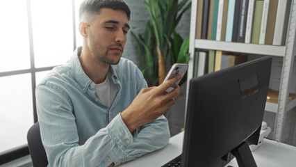 Young man using smartphone in modern office setting with computer and bookshelf in background, showcasing focused professional environment during work hours.
