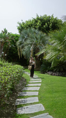 Woman walking along stone path in lush tropical resort garden with vibrant palm trees and exotic...