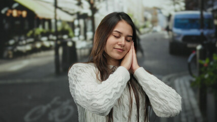 Young woman wearing a cozy sweater stands smiling and relaxed with hands together as if sleeping on a street with soft light and an out-of-focus urban background.