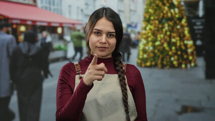 Young woman in apron pointing on street near cafe with festive lights and people walking in...