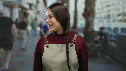 Hispanic woman waitress in apron smiling outdoors on city street with blurred background of people...