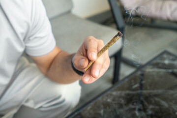 Man is holding a lit cannabis joint, exhaling smoke while seated on a couch in a contemporary living room. The ambiance feels relaxed and casual. 