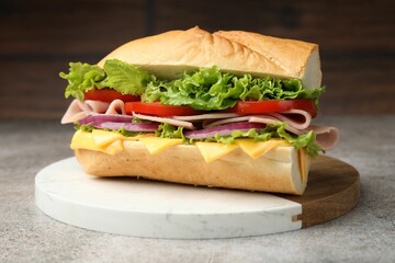 Tasty baguette sandwich with ham, cheese and vegetables on grey textured table against blurred...
