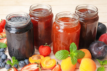 Tasty sweet jams in jars surrounded by ingredients on light wooden table, closeup