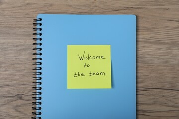 Sticky note with phrase Welcome To The Team and notebook on wooden table, top view