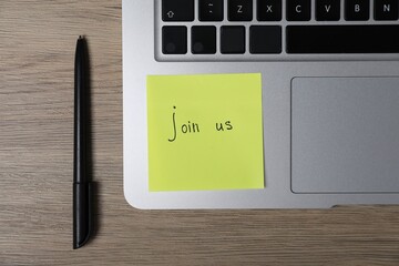 Sticky note with phrase Join Us, laptop and marker on wooden table, flat lay