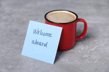 Sticky note with phrase Welcome Aboard and coffee on grey table, closeup