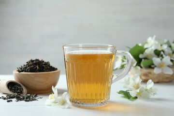 Aromatic jasmine tea in glass cup, flowers and dried leaves on white wooden table, closeup