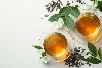 Aromatic jasmine tea in glass cups, flowers and leaves on white table, flat lay. Space for text