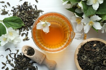 Aromatic jasmine tea in glass cup, flowers and leaves on white wooden table, flat lay