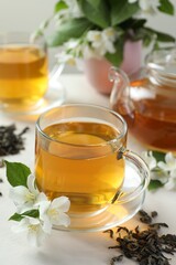 Aromatic jasmine tea in glass cups, teapot, flowers and dried leaves on white table, closeup