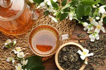 Aromatic jasmine tea in glass cup, teapot, brew and flowers on wooden table, flat lay