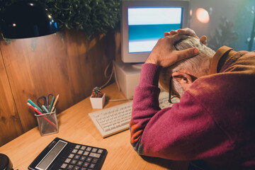 Elderly man frustratedly working at computer in home office with vintage equipment and ambient light