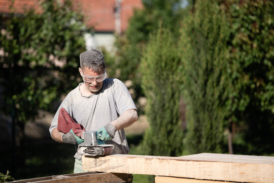 Carpenter sanding wooden plank outdoors with belt sander