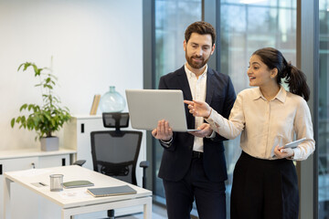 A businessman and businesswoman collaborate on a laptop in a modern office setting, working...