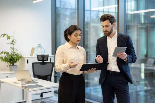 Two business professionals, a woman and a man, are collaborating in a modern office setting, reviewing documents and using a tablet.