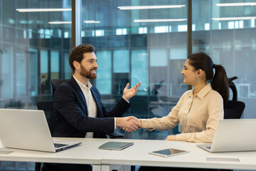 A man and a woman shake hands in an office setting, likely after a successful business meeting or agreement.