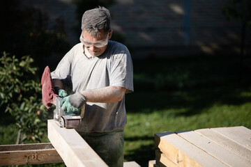 Carpenter sanding wooden plank outdoors with belt sander